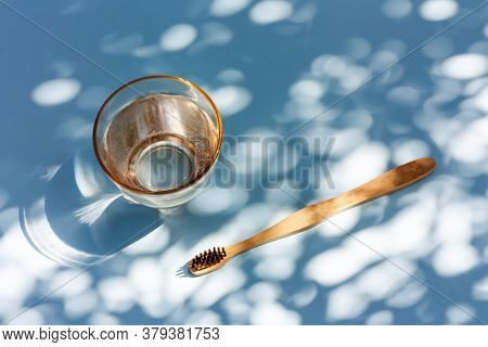 Bamboo Toothbrush And Glass Of Water On Light Blue Pastel Background With Shadows Of Natural Light. 