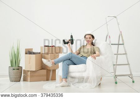 Portrait of smiling young woman in hardhat sitting with cordless screwdriver in armchair under cover in room with moving boxes