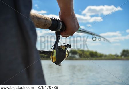 Fishing Rod Wheel Close-up In The Hands Of A Young Man On View Of River And Clouds