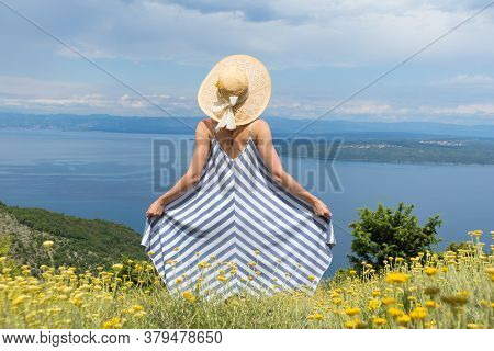 Rear View Of Young Woman Wearing Striped Summer Dress And Straw Hat Standing In Super Bloom Of Wildf