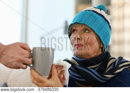 An Elderly Lady In A Knitted Hat Sits On A Sofa And Holds A Cup. Weak Immune System In Old Age. Cold