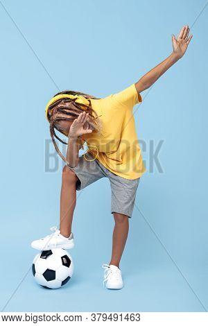 Cheerful Little Boy With African With Headphones Dabbing With One Leg On Soccer Ball Over Blue Backg
