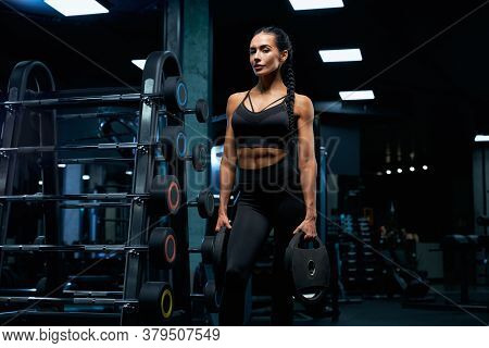 Side View Of Gorgeous Brunette Female Bodybuilder With Braids Posing Near Stand With Sports Equipmen
