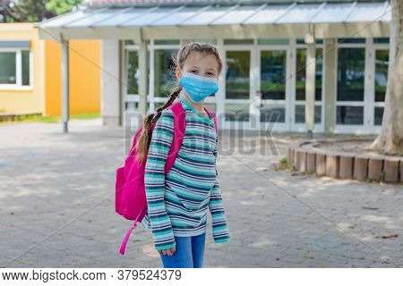 A Girl With A Crimson, Pink Backpack Stands In Front Of The School Doors In A Protective Mask. Back 