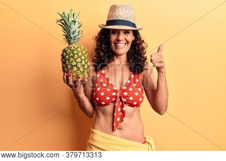 Middle age beautiful woman wearing bikini and summer hat holding pineappel smiling happy and positive, thumb up doing excellent and approval sign 