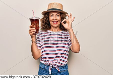 Middle age brunette woman on summer hat drinking fresh cola refeshment over isolated background doing ok sign with fingers, smiling friendly gesturing excellent symbol