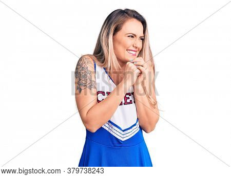 Young beautiful blonde woman wearing cheerleader uniform laughing nervous and excited with hands on chin looking to the side 