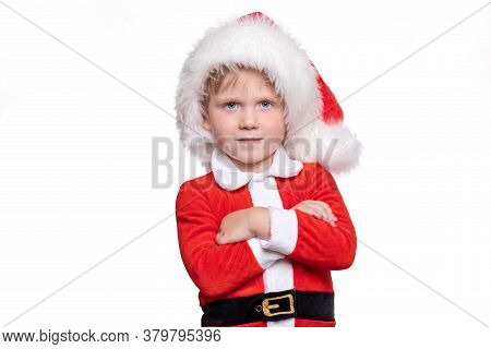 A Child Boy In A Red Suit And A Santa Claus Hat Stands With His Hands On His Chest. A Positive Story