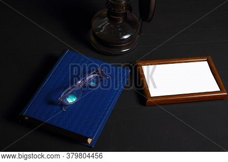 A Stack Of Old Books, A Oil Lamp, Empty Frame And Glasses On A Dark Background. Library At Night