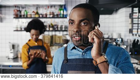 Close Up Of Male African American Barmen Talking On Mobile Phone In Cafe. Handsome Man Waiter Speaki