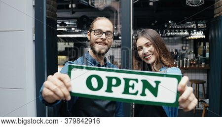 Portrait Of Caucasian Joyful Couple Of Bartenders In Aprons Smiling Happily To Camera And Holding Ta