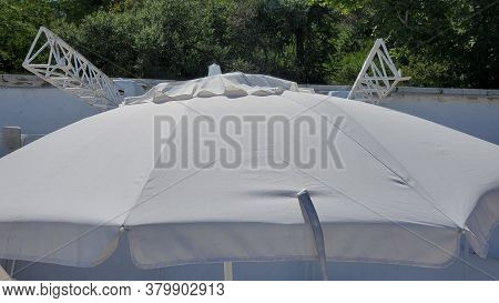 Big White Parasol At Beach With Green Trees On Background. Closeup Of Sunshade At Seaside. Summer Re