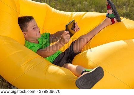 Cheerful Boy Lies In A Yellow Lamzack, Holding A Tablet In His Hands, Plays Or Communicates On Socia