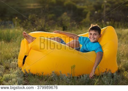 Cheerful Boy In A Blue T-shirt Lies On A Yellow Airy Sofa, On Green Grass, Outdoors