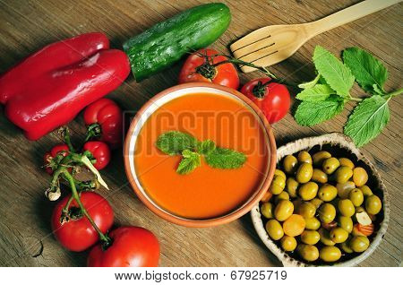 a bowl with spanish gazpacho, some vegetables to prepare it such as tomato, red pepper and cucumber, and a plate with gazpachas olives on a rustic wooden table