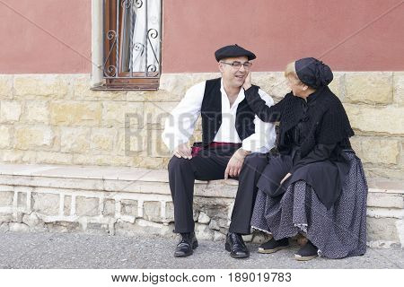 Couple dressed with one of the traditional Basque Country dresses outdoors seated in front of a house.