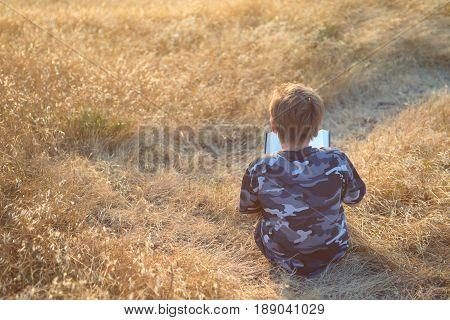 Boy reading book in yellow grass high in the hills, trail winding ahead of him, rear view