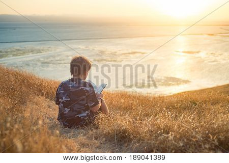 Boy reading book in yellow grass high in the hills, watching beautiful sunset above Bay Area, rear view
