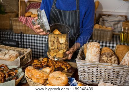 Mid section of staff holding glass jar of cookies at counter in bakery shop