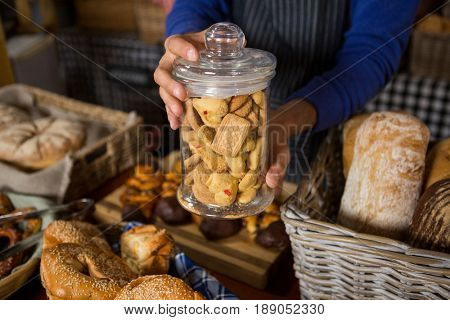 Mid section of staff holding glass jar of cookies at counter in bakery shop