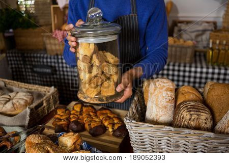 Mid section of staff holding glass jar of cookies at counter in bakery shop