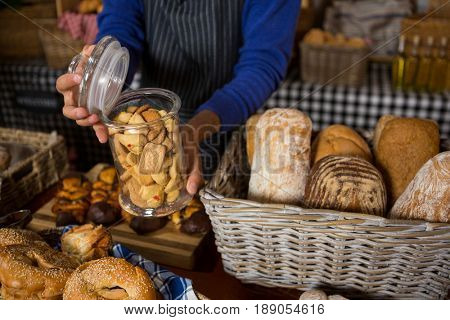 Mid section of staff holding glass jar of cookies at counter in bakery shop