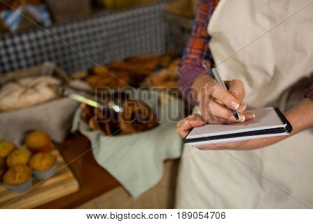 Mid section of female staff writing on notepad at bread counter in bakery shop