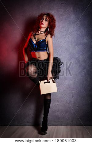 Curly redhead sexy girl looks like a doll holds a cardboard box on a gray background. poppet for adult men. Fashion style.