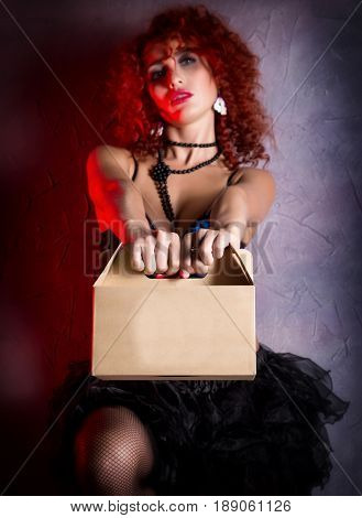 Curly redhead sexy girl looks like a doll holds a cardboard box on a gray background. poppet for adult men. Fashion style.