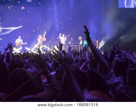 Audience with hands raised at a music festival and lights streaming down from above the stage. Focus on hands
