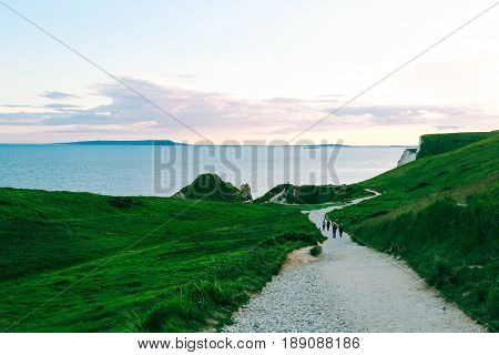 People walking on a path amongst greenery towards the famous Durdle Door on Jurassic Coast, Dorset, UK at dusk.