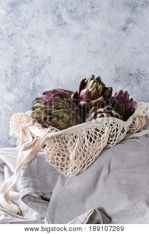 Uncooked whole organic wet purple artichokes in knitted bag on white tablecloth over gray texture background.