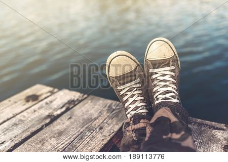 Man with crossed legs relaxing on wooden riverbank pier casual young guy wearing sneakers sitting by the river in summer afternoon selective focus
