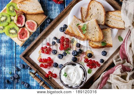 Variety of ingredients for making dessert sandwiches with bread, berries, cream cheese. Red currant, blueberries, sliced kiwi, figs over blue wood plank background. Flat lay, summer appetizer concept