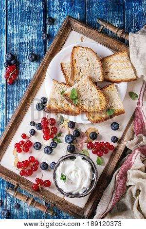 Variety of ingredients for making dessert sandwiches with bread, berries, cream cheese. Red currant, blueberries, sliced kiwi, figs over blue wood plank background. Flat lay, summer appetizer concept