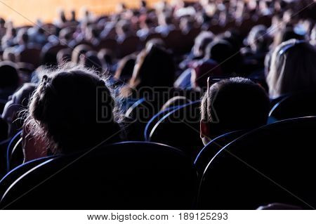 People, children, adults, parents in the theater watching the performance. People in the auditorium looking at the stage. Shooting from the back