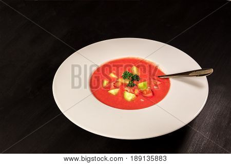 A plate of gazpacho soup with slices of tomato, cucumber, and onion, with parsley leaves, on a dark background with a place for text