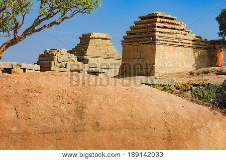 View of ancient ruins on Hemakuta hill in Hampi, Karnataka, India. Landscape with amazing stones tropical nature and temple under the blue sky. Monk stands near the ruins in orange robes