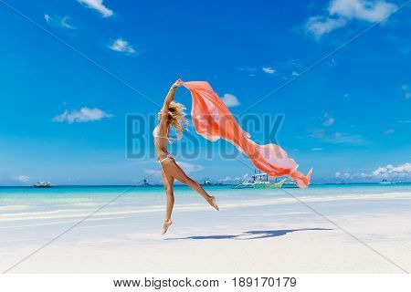 Happy beautiful young girl in pink bikini with waving in the wind pink fabric on a tropical beach. Blue sea in the background. Summer vacation concept.