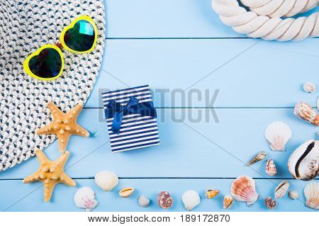 Hat and yellow sunglasses in shape of heart with shells starfishes gift box and sea rope on blue wooden background. Summer marine background. Beach accessories flat lay with negative space.