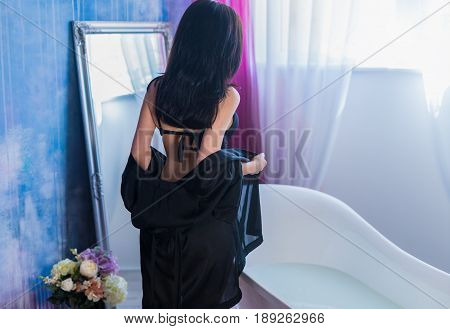 Sexy brunette woman taking off her clothes before bath. She is wearing black sexual lingerie