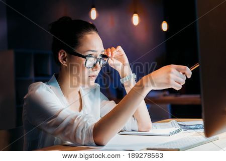 Concentrated Asian Businesswoman In Eyeglasses Pointing At Computer Monitor, Working Late At The Off