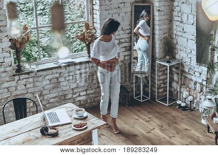 Fitting into skinny jeans. Full length of attractive young woman looking in the mirror while putting on her jeans
