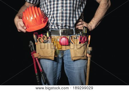 detail of handyman with leather toolsbelt and tools on dark background holding helmet