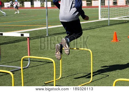 A high school male athlete bounds over yellow hurdles on a green turf field during practice