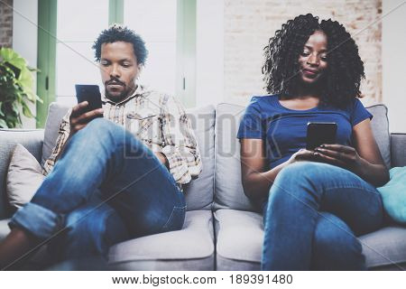 Upset african american couple relaxing together on the sofa.Young black man and his girlfriend using smartphones while rest at home in the living room. Horizontal, blurred background