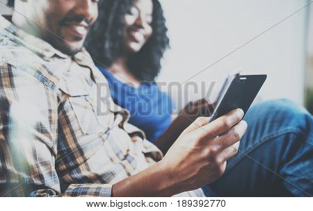 Smiling african american couple relaxing together on the couch.Young black man and his girlfriend using smartphones while rest at home in the living room.Horizontal, blurred background.Cropped