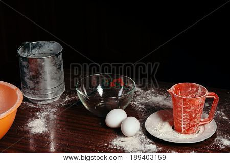 Orange cup measuring cup and a steel sieve two eggs stand on a wooden table on a black background. Flour.