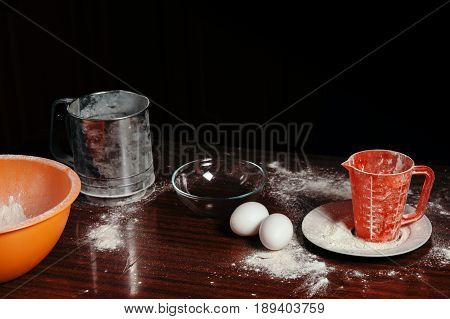 Orange cup measuring cup and a steel sieve two eggs stand on a wooden table on a black background. Flour.