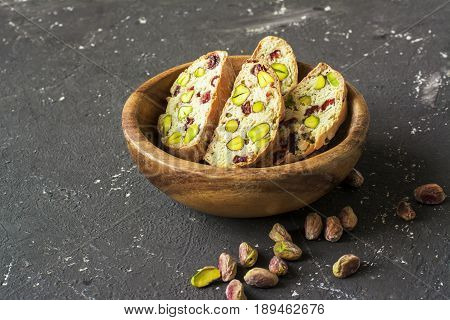 Traditional crispy Italian biscotti or cantuchini crackers with pistachios and dried cranberries in a bowl of olve wood on a dark background. Home classic healthy food